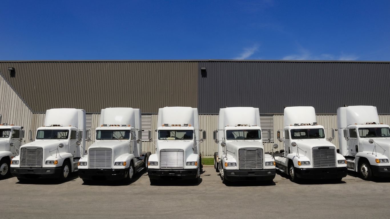 A line of white semi-trucks parked in front of a warehouse, representing fleet inventory tracked during fixed asset audits.